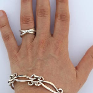 May include: A silver bracelet and ring set. The bracelet features an ornate design with curved links and a toggle clasp. The ring has a crisscross design. The jewelry is displayed on a hand against a white background.