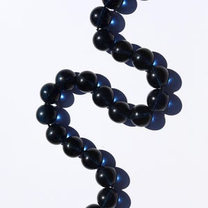 Op de afbeelding: Een ketting met grote, ronde, donkerblauwe kralen, geregen aan een dun, donker koord. De ketting heeft een zilverkleurige sluiting. Gepresenteerd op een witte achtergrond met schaduwen. De kralen zijn ongeveer 1 cm in diameter.