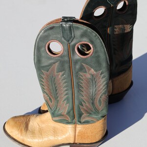 May include: A pair of tan leather cowboy boots with green leather tops. The boots have two decorative holes on each side and a brown leather pull tab on the back.