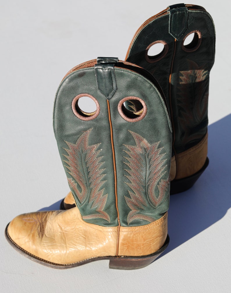 May include: A pair of tan leather cowboy boots with green leather tops. The boots have two decorative holes on each side and a brown leather pull tab on the back.