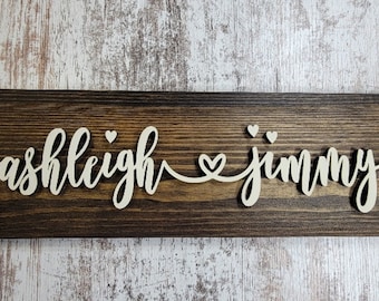 May include: A wooden sign with the names "ashleigh" and "jimmy" written in white cursive lettering with a heart symbol between the names.