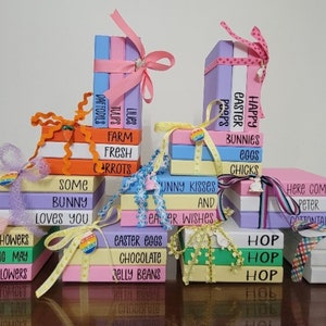 May include: A collection of stacked painted wooden books with Easter-themed sayings. The books are decorated with ribbons and bows in various colors, including pink, yellow, green, and blue. The sayings on the books include "Happy Easter Peeps", "Bunny Kisses and Easter Wishes", "Easter Eggs Chocolate Jelly Beans", "Hop Hop Hop", "Here Comes Peter Cottontail", and "Easter Blessings".