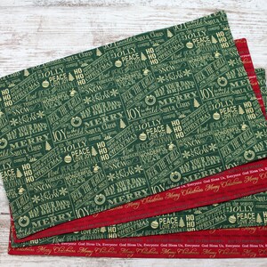 May include: Four green fabric placemats with a festive Christmas design. The placemats feature a repeating pattern of gold text on a dark green background. The text includes phrases like "Peace & Joy", "Merry Christmas", and "Deck the Halls".