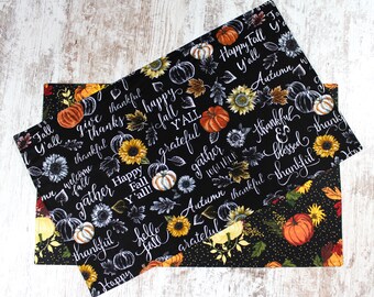 May include: Two black fabric placemats with a fall theme. The placemats feature a black background with white text and images of pumpkins, sunflowers, and leaves. The text includes phrases like "Happy Fall Y'all", "Gather Together", "Autumn", and "Thankful".