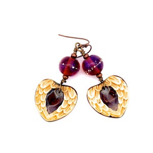 May include: A pair of dangle earrings with a heart-shaped design featuring a pair of wings. The heart is made of a cream-colored material with a brown glass bead in the center. The earrings are accented with a purple and brown glass bead.