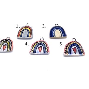 Five ceramic rainbow charms with different color schemes. Charm 1 is blue and yellow with a white background. Charm 2 is blue and white with a cream background. Charm 3 is red, yellow, and blue with a white background and a red heart. Charm 4 is yellow, blue, and red with a white background and a red heart. Charm 5 is yellow, blue, and red with a white background and a red heart. Each charm has a small metal loop for hanging.