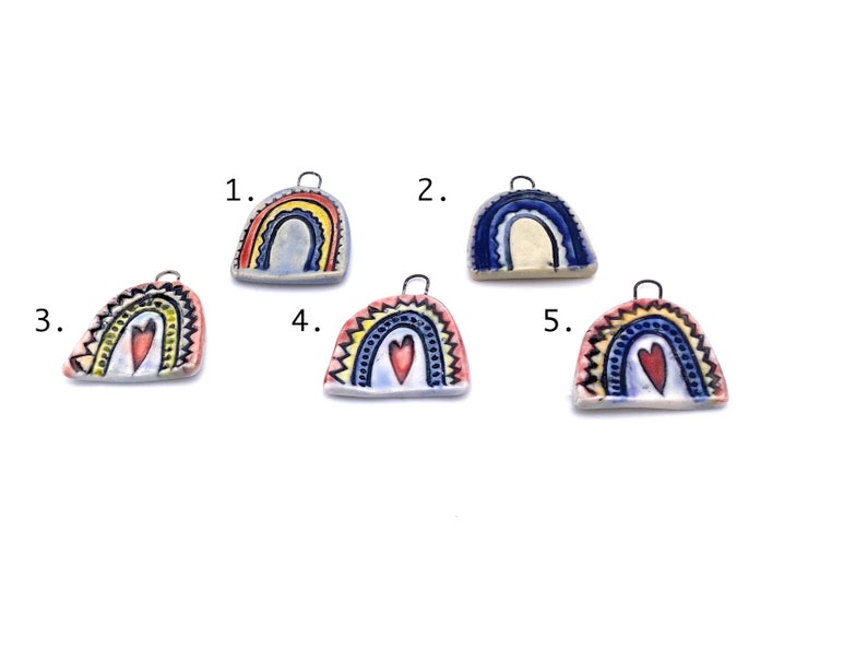 Five ceramic rainbow charms with different color schemes. Charm 1 is blue and yellow with a white background. Charm 2 is blue and white with a cream background. Charm 3 is red, yellow, and blue with a white background and a red heart. Charm 4 is yellow, blue, and red with a white background and a red heart. Charm 5 is yellow, blue, and red with a white background and a red heart. Each charm has a small metal loop for hanging.