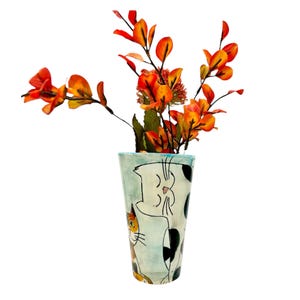 May include: A tall, hand-painted ceramic vase with a light blue background and cat illustrations. The vase holds orange and red artificial flowers. The vase has a unique, artistic design, perfect for home decor.