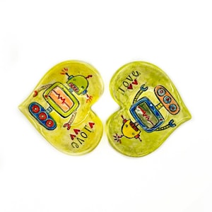 May include: Two heart-shaped ceramic dishes with a yellow-green hue. Each dish features a whimsical robot design with red, blue, and orange accents. The word "love" is written on each dish, adding a touch of affection.