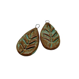 May include: Two ceramic leaf-shaped earrings. The earrings have a teardrop shape with a brown, textured exterior and a teal interior. The leaf design is carved out, revealing the teal color. Each earring has a small black wire loop at the top.