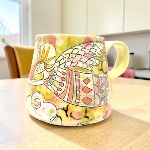 May include: A ceramic mug featuring a hand-painted illustration of an angel with a halo and wings. The design incorporates a vibrant color palette of yellow, pink, and blue with geometric patterns. The mug has a wide base and a curved handle.