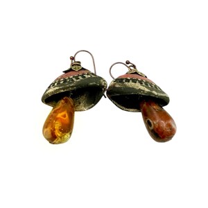 A pair of handmade ceramic and Red Jasper stone earrings lay on a white background with the tops facing away from the viewer. Each earring features a rustic handmade ceramic oblong-shaped bell in shades of dark brown and brick red with a red Jasper teardrop shaped bead dangling below. The earrings are hand wrapped with bronze wire and feature bronze ear wires.