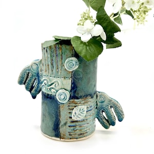May include: A blue and green ceramic vase with a unique, textured design. The vase has two small, wing-like handles on either side and features a patchwork of different patterns and colors.