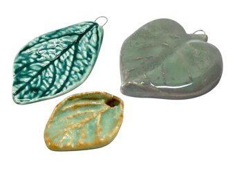 May include: Three ceramic leaf-shaped ornaments with glossy finishes. One is a dark teal elongated leaf, another is a rounded sage green leaf, and the third is a smaller, light mint green leaf with brown edges. Each piece features a small metal loop.