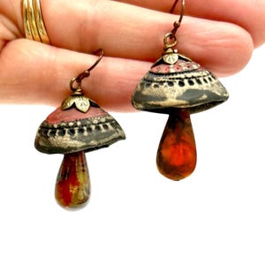 A pair of handmade ceramic and Red Jasper stone earrings hang from a human hand. Each earring features a rustic handmade ceramic oblong-shaped bell in shades of dark brown and brick red with a red Jasper teardrop shaped bead dangling below. The earrings are hand wrapped with bronze wire and feature bronze ear wires.
