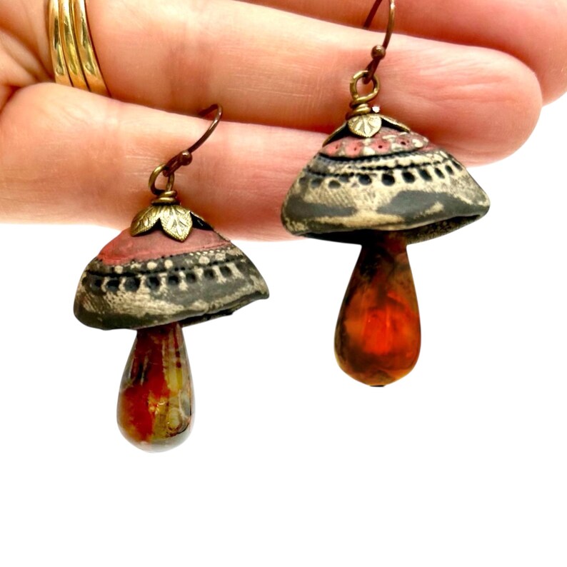 A pair of handmade ceramic and Red Jasper stone earrings hang from a human hand. Each earring features a rustic handmade ceramic oblong-shaped bell in shades of dark brown and brick red with a red Jasper teardrop shaped bead dangling below. The earrings are hand wrapped with bronze wire and feature bronze ear wires.