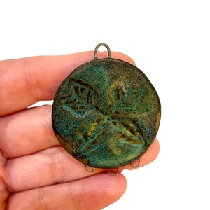 May include: Round, handcrafted pendant with a textured, teal and bronze finish. The pendant features a raised floral design in the center and a small loop at the top for hanging. The edges have small loops for attaching additional elements.