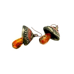 A pair of handmade ceramic and Red Jasper stone earrings lay on a white background with the tops towards the back, right. Each earring features a rustic handmade ceramic oblong-shaped bell in shades of dark brown and brick red with a red Jasper teardrop shaped bead dangling below. The earrings are hand wrapped with bronze wire and feature bronze ear wires.