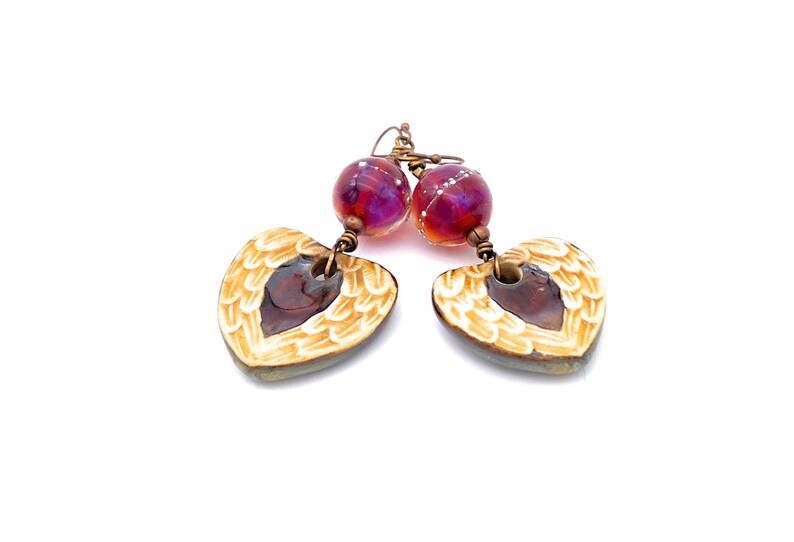 Earrings made with a heart-shaped charm featuring 2 angel wings in shades of cream with maroon in the center hang below matching maroon handmade lampwork glass beads.  Hand wrapped with copper wire.  Copper ear wires.