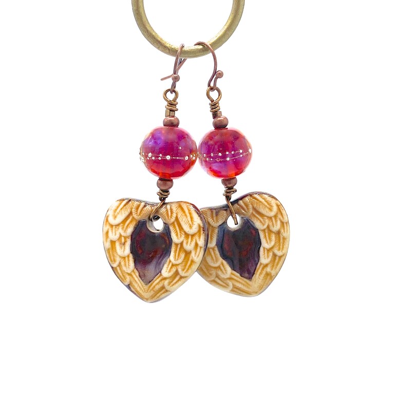 Earrings made with a heart-shaped charm featuring 2 angel wings in shades of cream with maroon in the center hang below matching maroon handmade lampwork glass beads.  Hand wrapped with copper wire.  Copper ear wires.