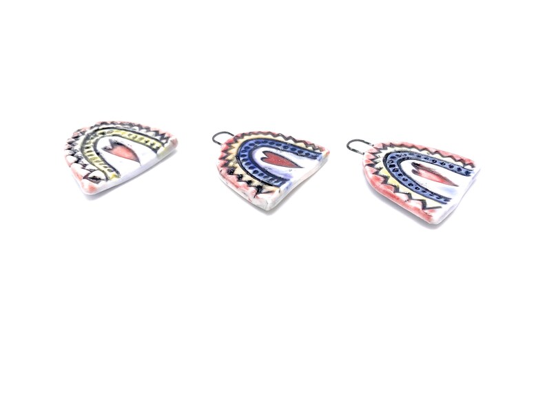 Three ceramic rainbow-shaped ornaments with a heart design in the center. The rainbows are painted in shades of blue, yellow, and red. The ornaments have a white background and a black outline.