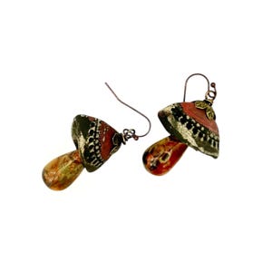 A pair of handmade ceramic and Red Jasper stone earrings lay on a white background with the tops towards the back, right. Each earring features a rustic handmade ceramic oblong-shaped bell in shades of dark brown and brick red with a red Jasper teardrop shaped bead dangling below. The earrings are hand wrapped with bronze wire and feature bronze ear wires.