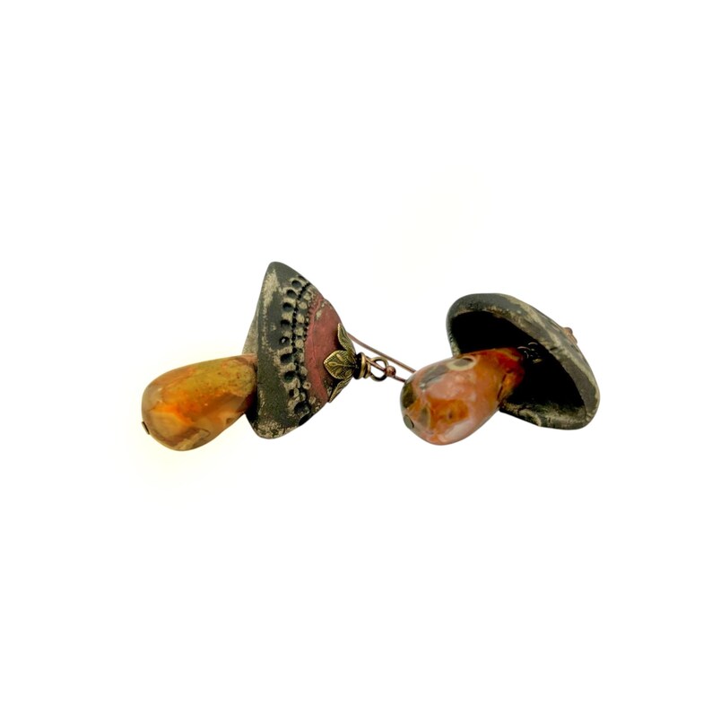 A pair of handmade ceramic and Red Jasper stone earrings lay on a white background with the tops towards the back, right. Each earring features a rustic handmade ceramic oblong-shaped bell in shades of dark brown and brick red with a red Jasper teardrop shaped bead dangling below. The earrings are hand wrapped with bronze wire and feature bronze ear wires.