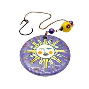May include: A ceramic sun-shaped ornament with a purple background and yellow sun rays. The sun has a smiling face with orange cheeks. It hangs from a chain with a hook and decorative beads. The ornament is approximately 3 inches in diameter.