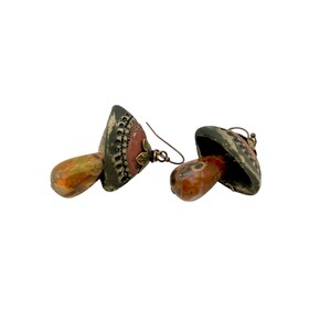 A pair of handmade ceramic and Red Jasper stone earrings lay on a white background with the tops towards the back, right. Each earring features a rustic handmade ceramic oblong-shaped bell in shades of dark brown and brick red with a red Jasper teardrop shaped bead dangling below. The earrings are hand wrapped with bronze wire and feature bronze ear wires.