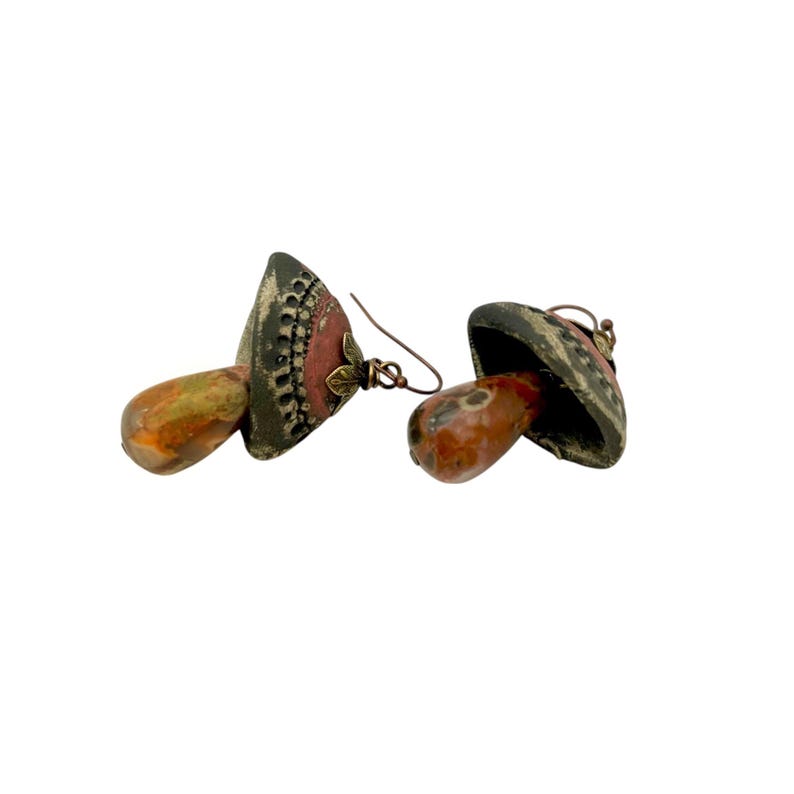 A pair of handmade ceramic and Red Jasper stone earrings lay on a white background with the tops towards the back, right. Each earring features a rustic handmade ceramic oblong-shaped bell in shades of dark brown and brick red with a red Jasper teardrop shaped bead dangling below. The earrings are hand wrapped with bronze wire and feature bronze ear wires.