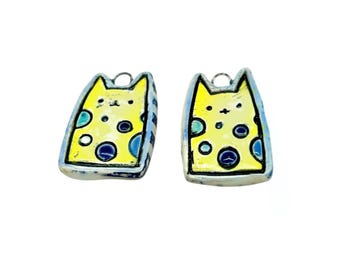 May include: Two cat-shaped earrings. Each earring features a yellow cat face with blue and green accents. The earrings have a silver-colored border and a small loop at the top for hanging. The cat's face has black dots for eyes and a nose.