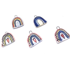 Five ceramic rainbow charms with a heart in the center. The rainbows are painted in various colors including blue, yellow, red, and white. The charms have a silver loop for hanging.