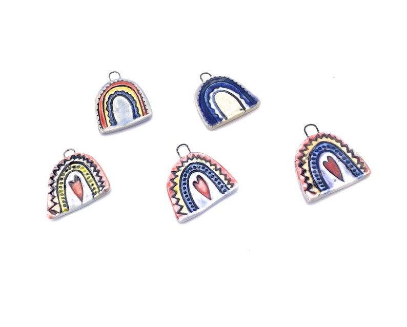 Five ceramic rainbow charms with a heart in the center. The rainbows are painted in various colors including blue, yellow, red, and white. The charms have a silver loop for hanging.