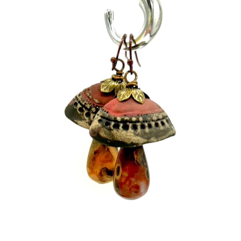 A pair of handmade ceramic and red jasper stone earrings hang from a silver hook against a white background. Each earring features a rustic handmade ceramic oblong-shaped bell in shades of dark brown and brick red with a red Jasper teardrop shaped bead dangling below. The earrings are hand wrapped with bronze wire and feature bronze ear wires.