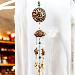 May include: A decorative wind chime featuring a sun, cloud, star, and crescent moon design. The sun and cloud are brown, with green and white beads. Three gold-colored bells hang below. A unique, handcrafted home decor item.