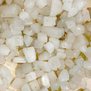 May include: A collection of tumbled, milky white gemstones. The stones vary in size and shape, with rounded edges and a smooth, polished appearance. The gemstones are scattered across a light-colored surface, creating a natural, organic aesthetic.