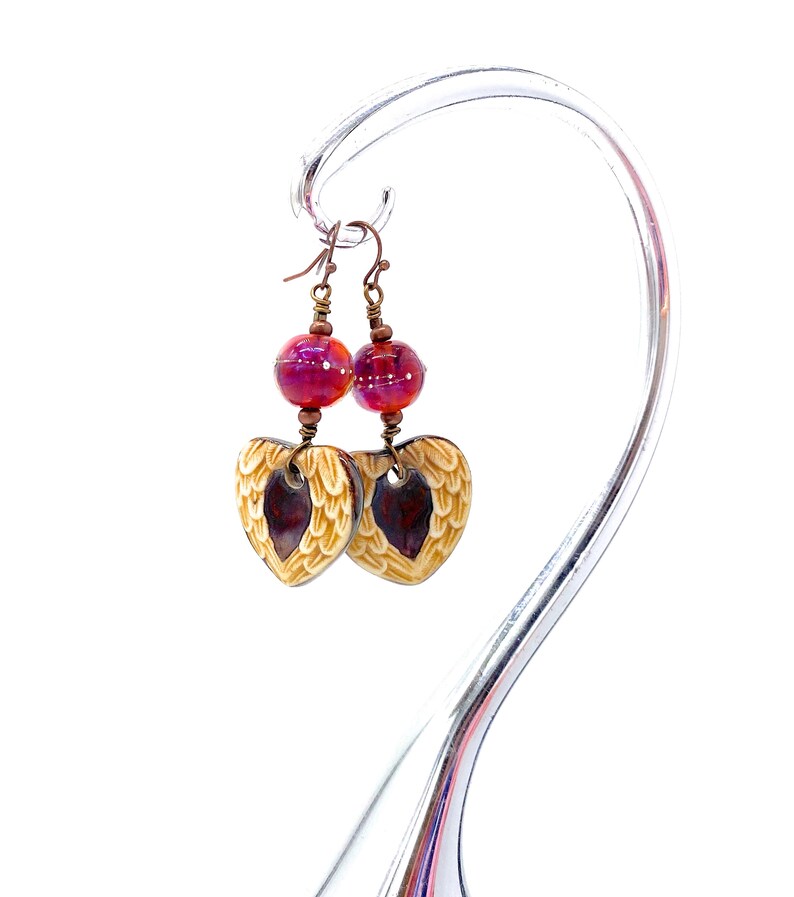 Earrings made with a heart-shaped charm featuring 2 angel wings in shades of cream with maroon in the center hang below matching maroon handmade lampwork glass beads.  Hand wrapped with copper wire.  Copper ear wires.