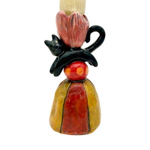 May include: A whimsical ceramic candlestick holder featuring a black cat perched atop a red and yellow base. The candlestick has a red flower-shaped top and is painted with vibrant colors. A unique decorative piece.
