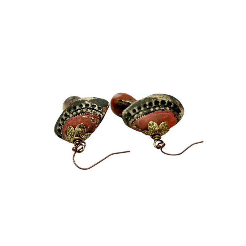 A pair of handmade ceramic and Red Jasper stone earrings lay on a white background with the tops towards the viewer. Each earring features a rustic handmade ceramic oblong-shaped bell in shades of dark brown and brick red with a red Jasper teardrop shaped bead dangling below. The earrings are hand wrapped with bronze wire and feature bronze ear wires.