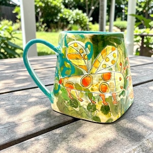 May include: A square-shaped ceramic mug with a teal handle and rim. The exterior features a hand-painted design of a yellow and orange insect with wings against a green and yellow mottled background. The mug has a wide base and a smooth finish.