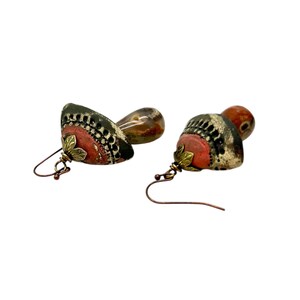 A pair of handmade ceramic and Red Jasper stone earrings lay on a white background with the tops towards the front, left. Each earring features a rustic handmade ceramic oblong-shaped bell in shades of dark brown and brick red with a red Jasper teardrop shaped bead dangling below. The earrings are hand wrapped with bronze wire and feature bronze ear wires.