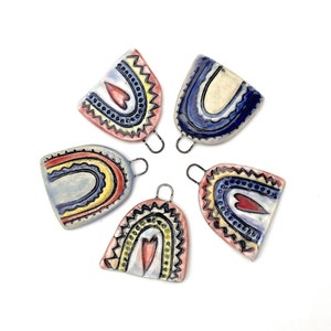 Five ceramic rainbow charms with heart details. The charms are in various colors including blue, red, yellow, and white. Each charm has a small loop at the top for hanging.