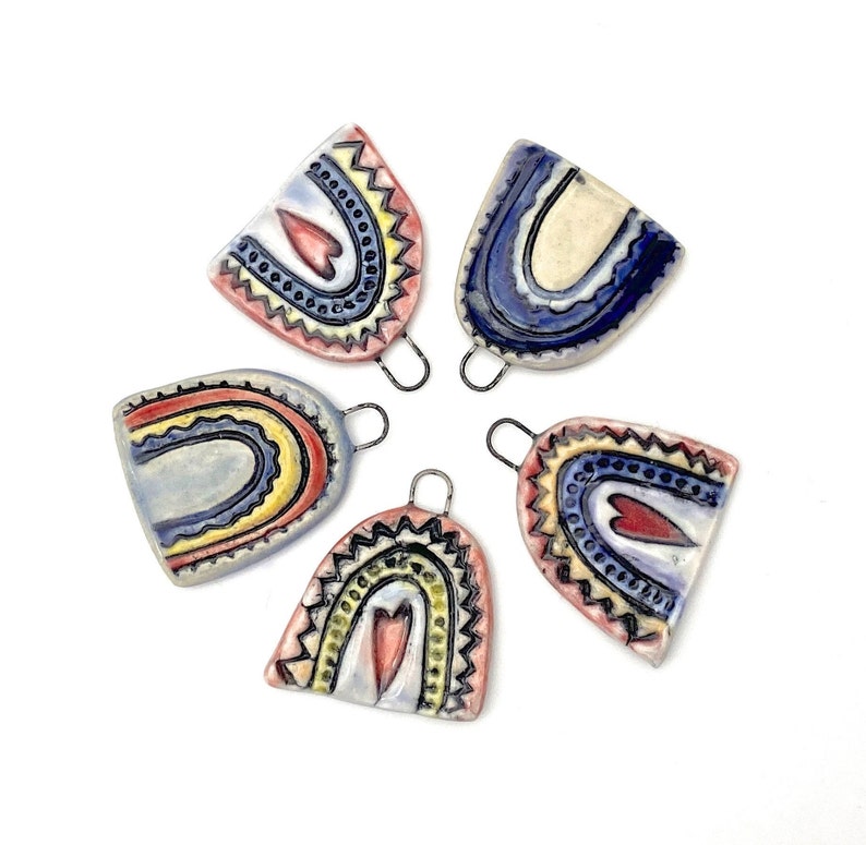 Five ceramic rainbow charms with heart details. The charms are in various colors including blue, red, yellow, and white. Each charm has a small loop at the top for hanging.
