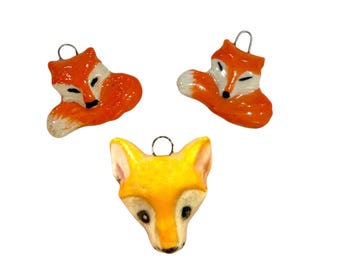 May include: Three fox-shaped charms with glossy finishes. Two charms depict curled foxes in vibrant orange with white accents and black details. The third charm is a yellow fox head with pointed ears, black eyes, and a small metal loop for hanging.