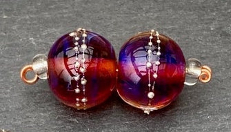 View of the 2 translucent maroon colored handmade lampwork glass beads with silver dots around the middle.