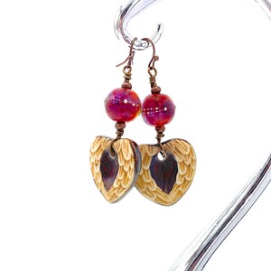 Earrings made with a heart-shaped charm featuring 2 angel wings in shades of cream with maroon in the center hang below matching maroon handmade lampwork glass beads.  Hand wrapped with copper wire.  Copper ear wires.