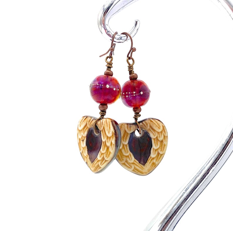 Earrings made with a heart-shaped charm featuring 2 angel wings in shades of cream with maroon in the center hang below matching maroon handmade lampwork glass beads.  Hand wrapped with copper wire.  Copper ear wires.