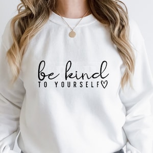 Pode incluir: Camisola branca com texto preto que diz "seja gentil consigo mesmo" com um pequeno símbolo de coração.