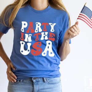 May include: A woman wearing a blue t-shirt with the text "Party in the USA" in red, white, and blue letters. She is holding a small American flag in her right hand.