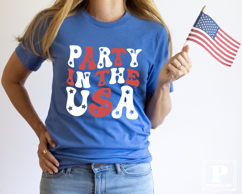 May include: A woman wearing a blue t-shirt with the text "Party in the USA" in red, white, and blue letters. She is holding a small American flag in her right hand.
